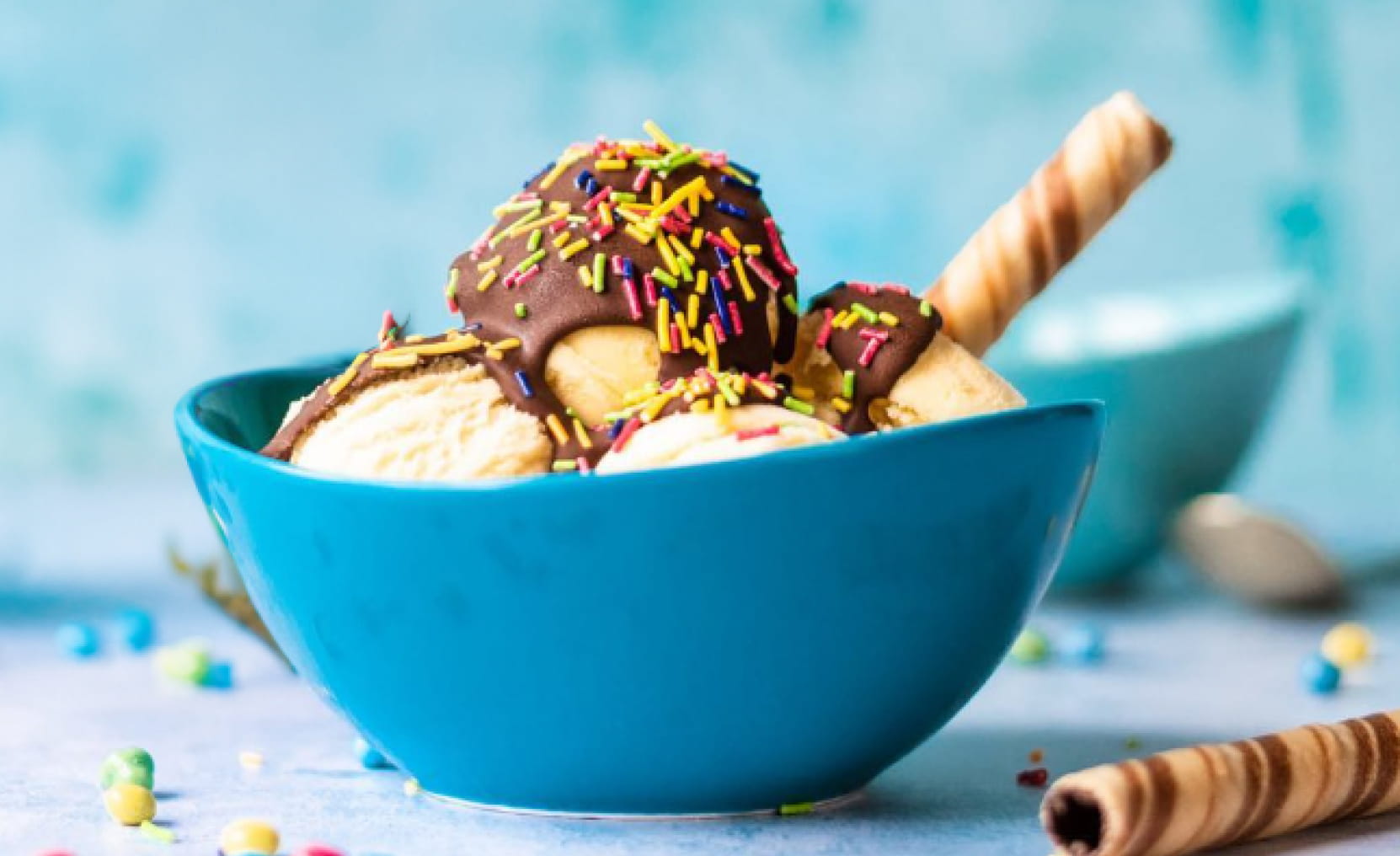 A colourful bowl of sprinkle topped vanilla ice cream covered in Askeys Crackin chocolate sauce sits in front of a bottle.