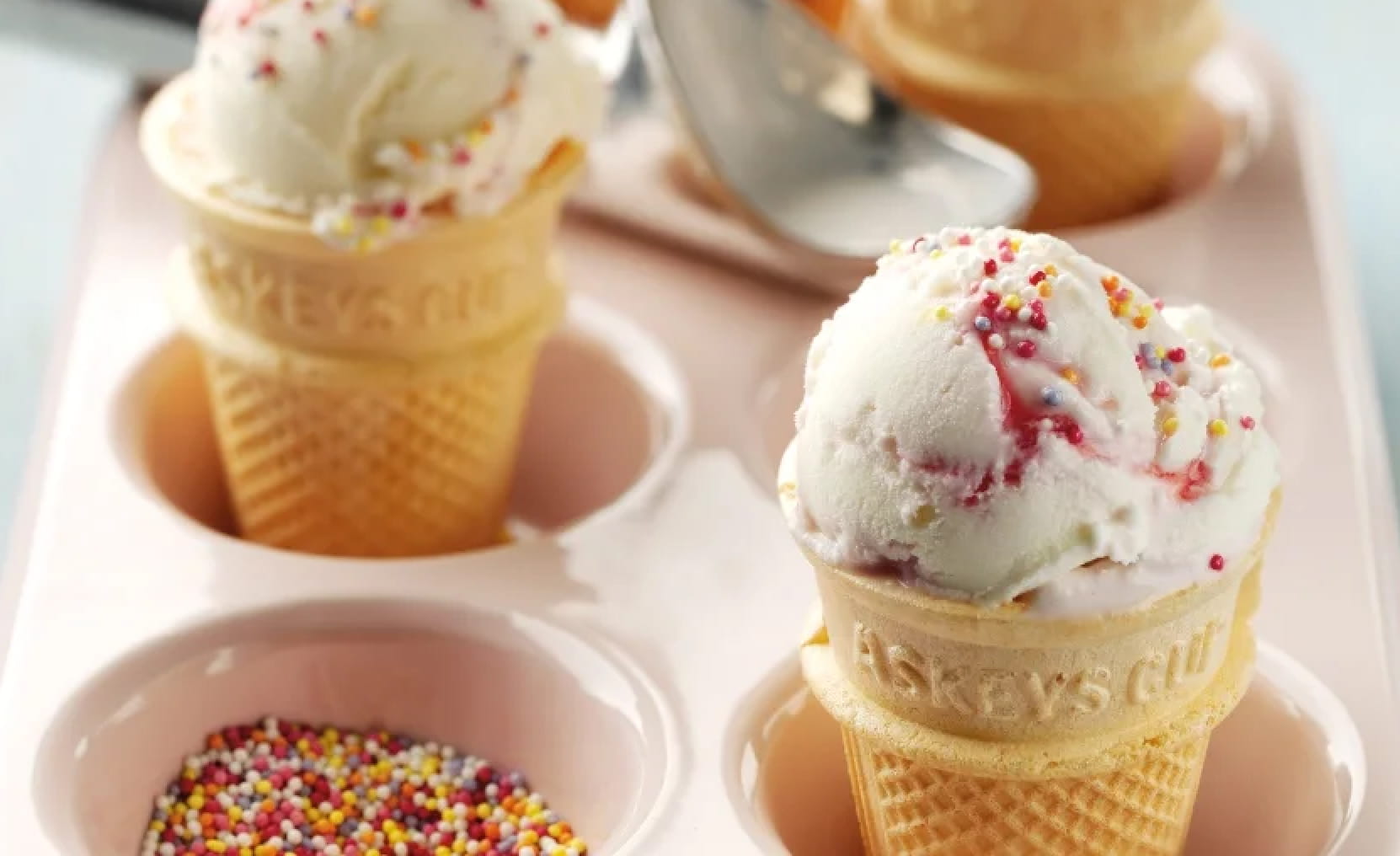 Wafer cones filled with different ice cream flavours and topped with Askeys sauce and sprinkles on a wooden serving board. Behind them, Askeys sauces for ice cream in all their different flavours.