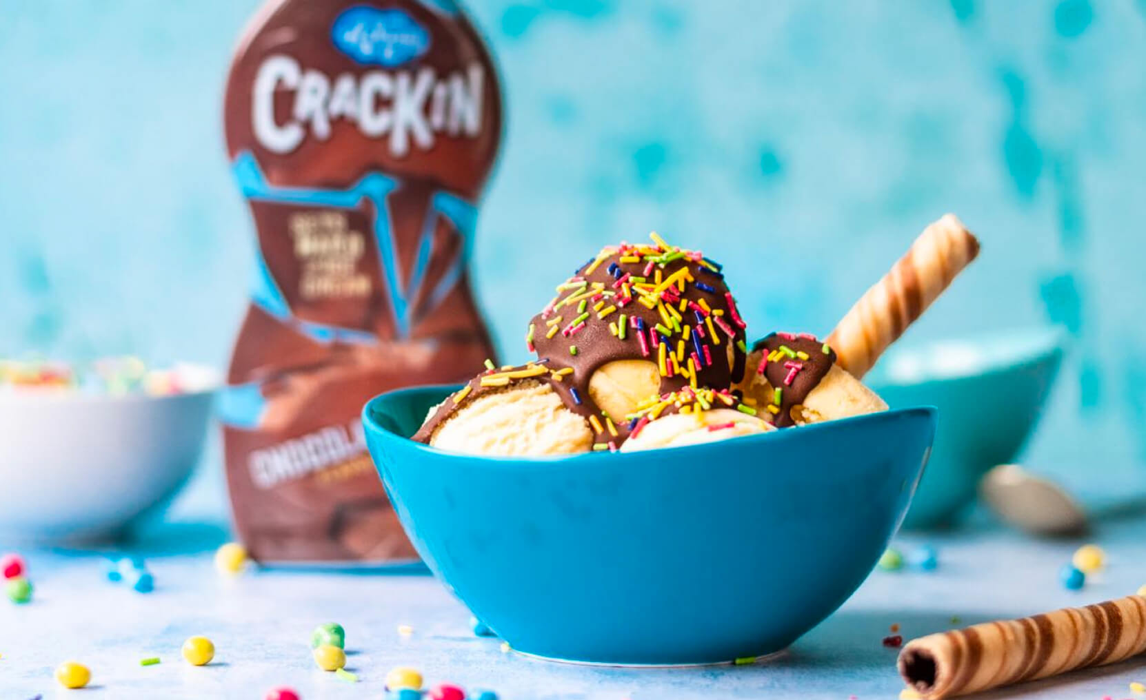 A colourful bowl of sprinkle topped vanilla ice cream covered in Askeys Crackin chocolate sauce sits in front of a bottle.