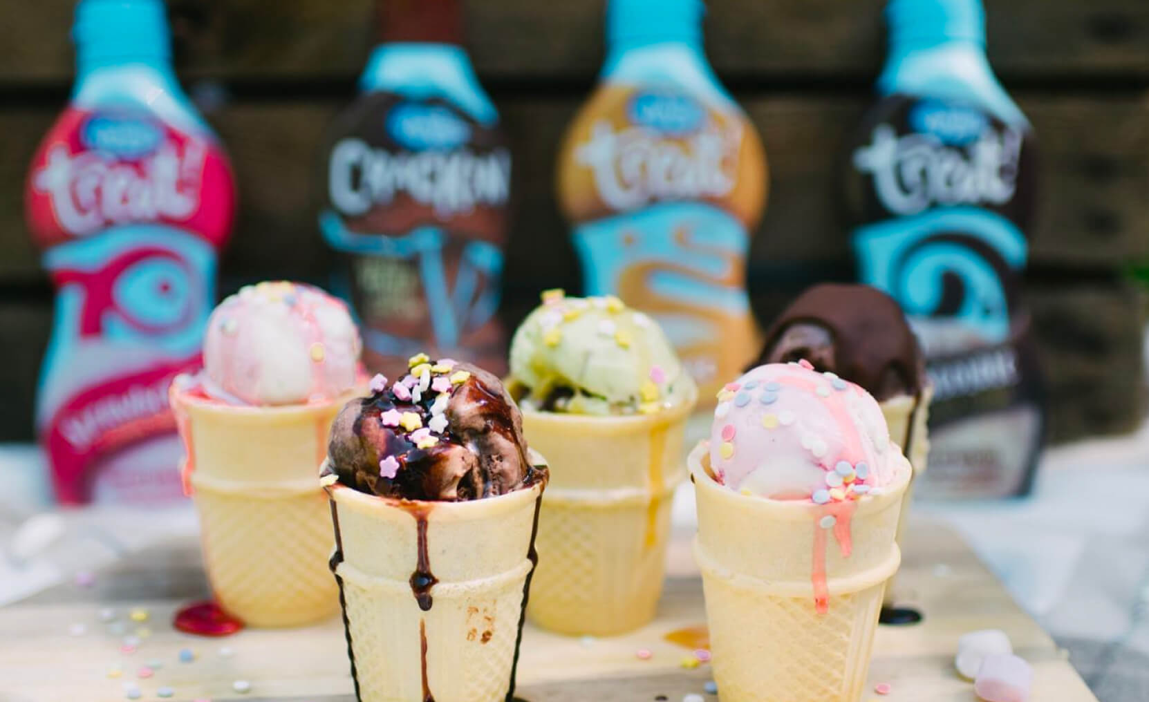 Wafer cones filled with different ice cream flavours and topped with Askeys sauce and sprinkles on a wooden serving board. Behind them, Askeys sauces for ice cream in all their different flavours.